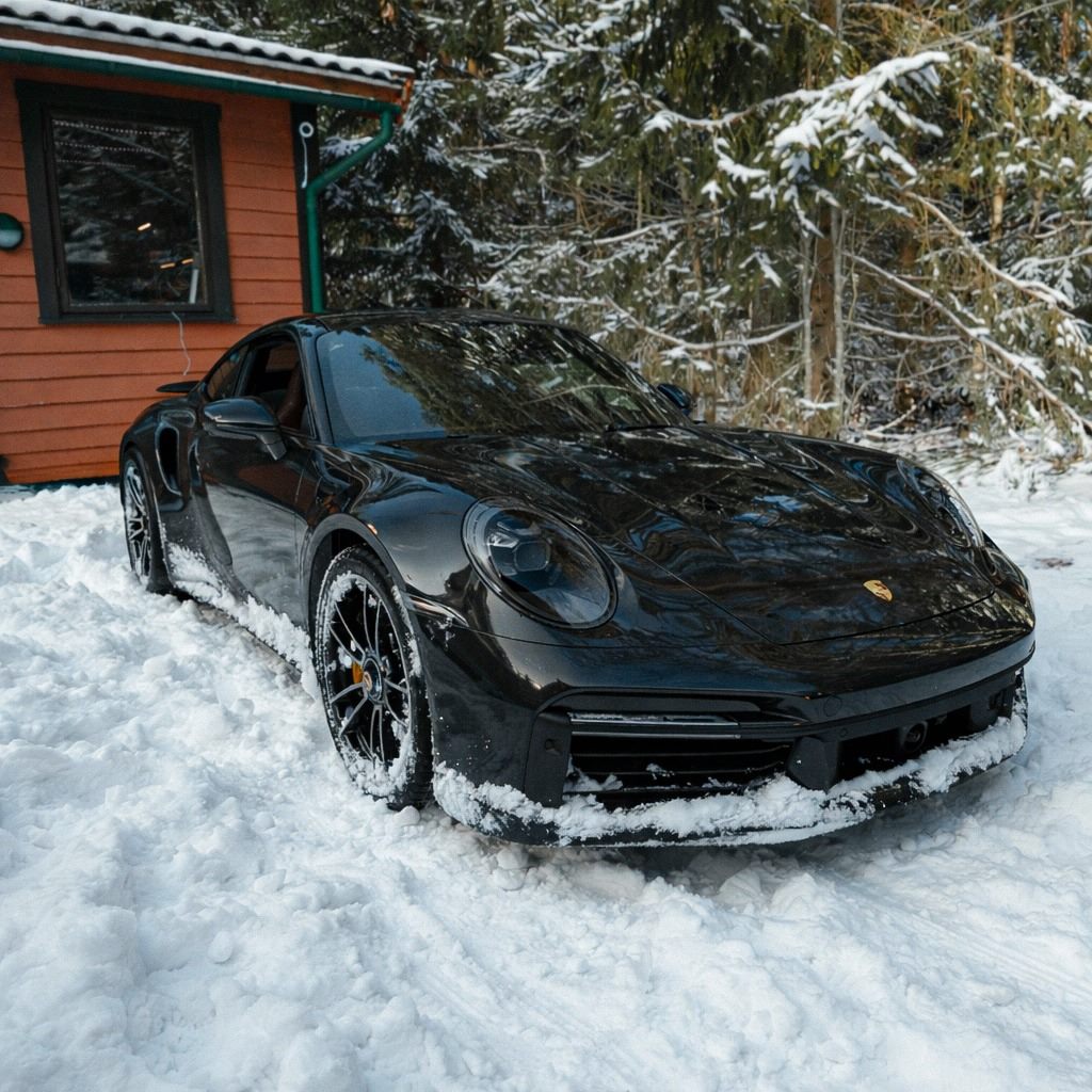 Black Porsche 911 parked in snow beside a red wooden building and snowy trees