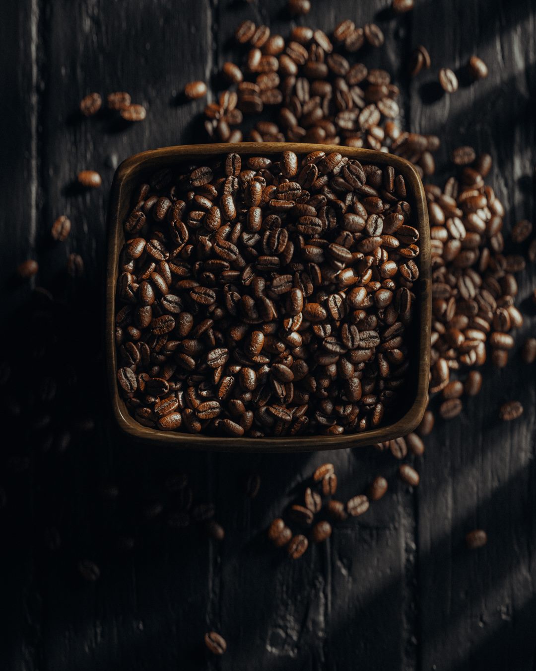 Coffee beans in and around a square bowl on dark wood